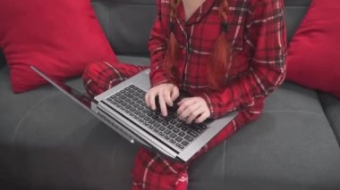 Evdeki kanepede dizüstü bilgisayarı, Noel ağacı ve hediyeleri olan kırmızı elbiseli bir kadın. Noel Baba şapkalı bir kız bilgisayarda video aracılığıyla iletişim kuruyor. İnternetten öğreniyorum. Yeni yıl alışverişi ve siparişi
