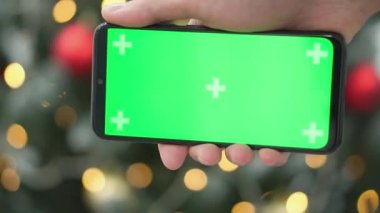 Kroma tuşuna yeşil ekran takılı akıllı telefon. Noel arkaplanındaki yeni yıl şenlikli yılbaşı ağacı arka planda bulanık ışıklar. Terfi, online alışveriş, iş.