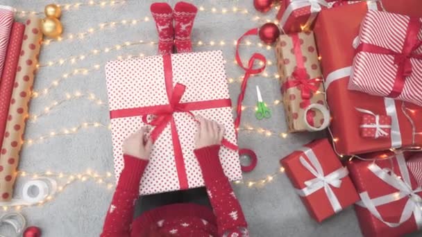 背景クリスマスライト ガーランド 暖炉のクリスマスギフトボックスの近くの女の子 若い女性は大晦日に木の近くに赤い紙で贈り物を包みます 雰囲気のお祝いムード 装飾冬 — ストック動画