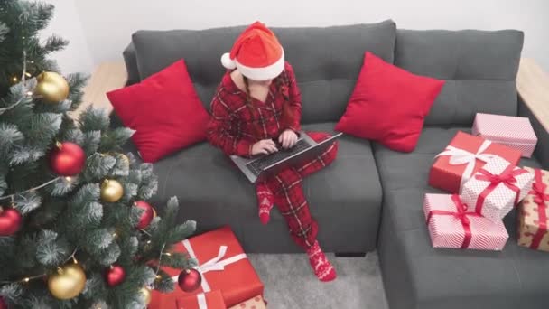 ノートパソコン クリスマスツリーと贈り物を自宅でソファの上に赤の女性 サンタクロースの帽子の女の子は コンピュータ上のビデオ通話を介して通信します オンラインインターネットを学ぶ 年末年始の買い物 — ストック動画