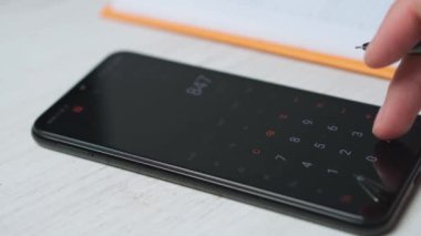 Genç bir girişimci telefonda hesaplamalar yapar ve verileri bir deftere yazar. Telefondaki hesap makinesini kullanan bir adamın yakın çekimi. Paranın hesaplanması, ödemeler, gelir, kar.