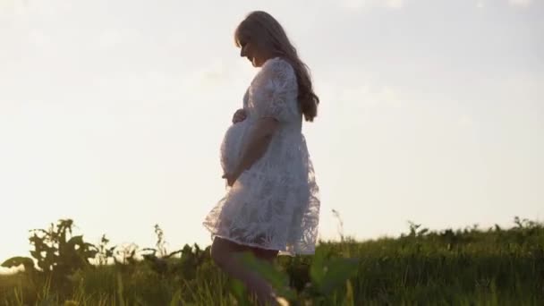 若い妊婦さんが夏に自然の中を歩いています 白いドレスを着た女性 妊娠中の女性が子供の誕生を待っています 未来の若い母親は赤ちゃんの誕生を待っています — ストック動画
