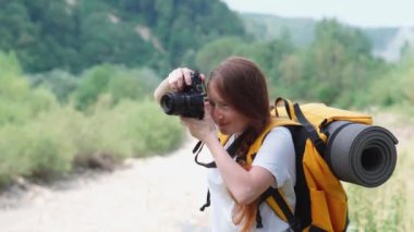 Kamera, turuncu sırt çantası ve şapkayla yürüyüş. Bir turist kız gezi sırasında doğanın fotoğraflarını çeker. Genç bir kadın DSLR kamerasıyla fotoğraf çekiyor. Ormanda ve dağlarda dinlenmek için bir yolculuk..