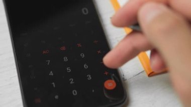 Genç bir girişimci telefonda hesaplamalar yapar ve verileri bir deftere yazar. Telefondaki hesap makinesini kullanan bir adamın yakın çekimi. Paranın hesaplanması, ödemeler, gelir, kar.