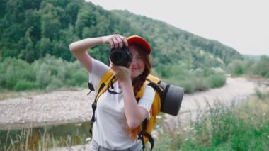 Kamera, turuncu sırt çantası ve şapkayla yürüyüş. Bir turist kız gezi sırasında doğanın fotoğraflarını çeker. Genç bir kadın DSLR kamerasıyla fotoğraf çekiyor. Ormanda ve dağlarda dinlenmek için bir yolculuk..