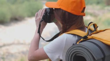 Kamera, turuncu sırt çantası ve şapkayla yürüyüş. Bir turist kız gezi sırasında doğanın fotoğraflarını çeker. Genç bir kadın DSLR kamerasıyla fotoğraf çekiyor. Ormanda ve dağlarda dinlenmek için bir yolculuk..