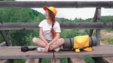 Dişi turist bir köprü üzerinde sırt çantasıyla oturur ve bekler. Turuncu sırt çantalı, şapkalı ve kameralı bir kız. Aktif ve turist yaşam tarzı kavramı.