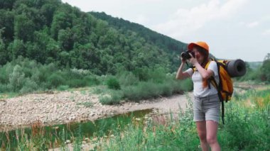 Kamera, turuncu sırt çantası ve şapkayla yürüyüş. Bir turist kız gezi sırasında doğanın fotoğraflarını çeker. Genç bir kadın DSLR kamerasıyla fotoğraf çekiyor. Ormanda ve dağlarda dinlenmek için bir yolculuk..