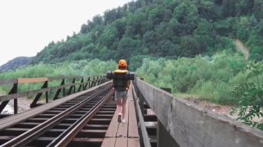 Turuncu sırt çantalı, şapkalı ve kameralı bir kız. Bir kadın turist, köprüdeki tren yolu boyunca sırt çantasıyla yürüyor. Aktif ve turist yaşam tarzı kavramı.