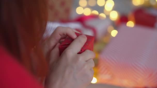 戴着圣诞礼帽的女孩用红色包装纸包装新年礼物 年轻妇女正在为她的朋友和亲戚准备圣诞礼物 准备过冬假期 — 图库视频影像