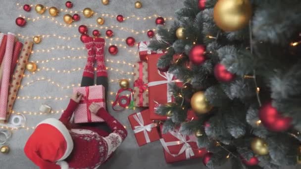背景クリスマスライト ガーランド 暖炉のクリスマスギフトボックスの近くの女の子 若い女性は大晦日に木の近くに赤い紙で贈り物を包みます 雰囲気のお祝いムード 装飾冬 — ストック動画