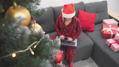 Evdeki kanepede dizüstü bilgisayarı, Noel ağacı ve hediyeleri olan kırmızı elbiseli bir kadın. Yeni yıl alışverişi ve sipariş. Noel Baba şapkalı bir kız bilgisayarda video aracılığıyla iletişim kuruyor. İnternet üzerinden öğrenme