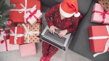Evdeki kanepede dizüstü bilgisayarı, Noel ağacı ve hediyeleri olan kırmızı elbiseli bir kadın. Yeni yıl alışverişi ve sipariş. Noel Baba şapkalı bir kız bilgisayarda video aracılığıyla iletişim kuruyor. İnternet üzerinden öğrenme
