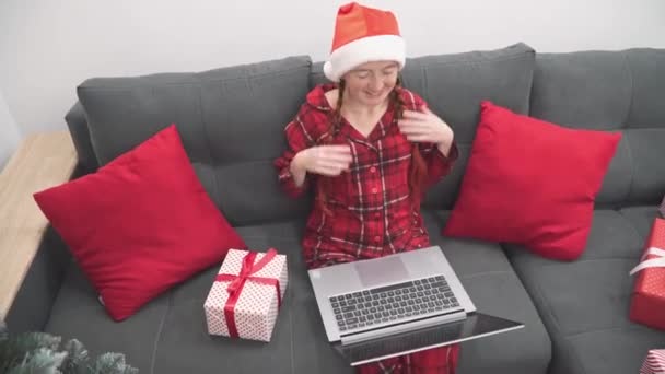 ノートパソコン クリスマスツリーと贈り物を自宅でソファの上に赤の女性 新年の休日のショッピングと注文 サンタクロースの帽子の女の子は コンピュータ上のビデオ通話を介して通信します オンラインインターネットを学ぶ — ストック動画