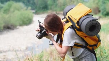 Genç bir kadın DSLR kamerasıyla fotoğraf çekiyor. Kamera, turuncu sırt çantası ve şapkayla yürüyüş. Bir turist kız gezi sırasında doğanın fotoğraflarını çeker. Ormanda ve dağlarda dinlenmek için bir yolculuk..