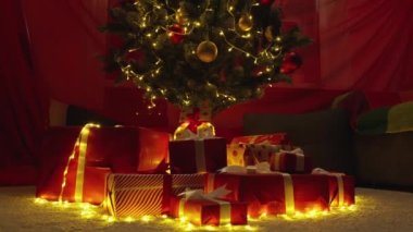 Noel ağacında ışıklar, toplar ve hediyelerle süslenmiş. Yılbaşı tatili atmosferi, kış masalı ve evde Noel ruhu. Yeşil ağacın altında kurdeleli kırmızı paketler. Yavaş çekim.