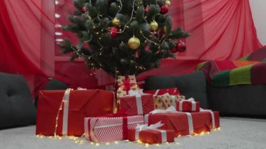 Noel ağacında ışıklar, toplar ve hediyelerle süslenmiş. Yılbaşı tatili atmosferi, kış masalı ve evde Noel ruhu. Yeşil ağacın altında kurdeleli kırmızı paketler. Yavaş çekim.