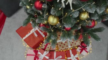 Noel ağacında ışıklar, toplar ve hediyelerle süslenmiş. Yılbaşı tatili atmosferi, kış masalı ve evde Noel ruhu. Yeşil ağacın altında kurdeleli kırmızı paketler. Yavaş çekim.