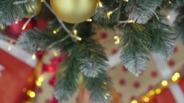Noel ağacında ışıklar, toplar ve hediyelerle süslenmiş. Yılbaşı tatili atmosferi, kış masalı ve evde Noel ruhu. Yeşil ağacın altında kurdeleli kırmızı paketler. Yavaş çekim.
