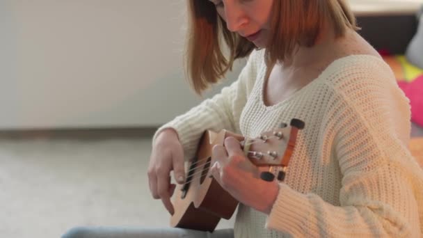 ウクレレを女性の手に ソファに座りながら家でウクレレを弾くことを学ぶ女性 女の子は小さなギターと歌でウクレレを演奏します 楽器の弦コンセプト — ストック動画