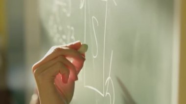 A girl student writes on a green board. Female teacher near the school board. A hand writes information on the board with white chalk. Concept of educational process, demonstration.