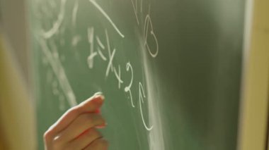 A girl student writes on a green board. Female teacher near the school board. A hand writes information on the board with white chalk. Concept of educational process, demonstration.