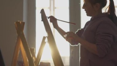A talented, innovative female artist paints with a brush on a canvas against the background of the sunset shining through window at home. A modern artist creates abstract modern art. The girl draws