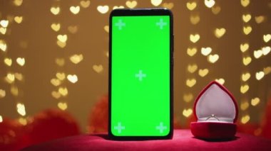 Sevgililer Günü için krom anahtarlı yeşil ekran akıllı telefon. Kırmızı arka plan, odaklı olmayan ışıklar, kalpler. Kırmızı mücevher kutusunda nişan yüzüğü ve evlilik teklifi konsepti. Tatil için online alışveriş