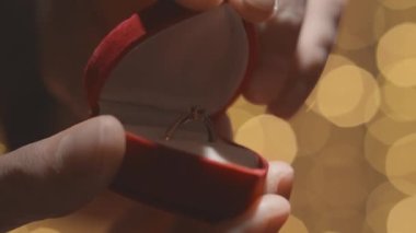 Alyans ve mücevher için kırmızı kutuyu açan elleri yakın plan. Sevgililer Günü ve evlilik teklifi kalplerin arka planında. Nişan, erkek kadına pırlantalı altın bir nişan yüzüğü verir.