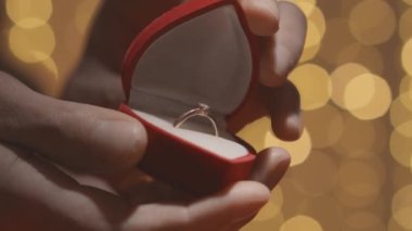 Close-up of hands opening a red box for a wedding ring and jewelry. Valentines Day and marriage proposal on the background of hearts. Engagement, man gives woman a gold engagement ring with a diamond
