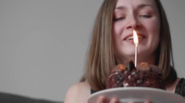 A young woman is celebrating her birthday alone. A girl at home with a cake lights candle, makes wish and blows out candle. In black and red clothes, on gray background, holiday celebrates anniversary