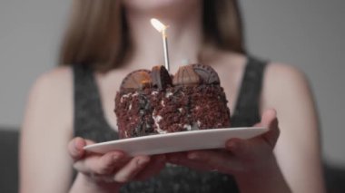 A young woman is celebrating her birthday alone. A girl at home with a cake lights candle, makes wish and blows out candle. In black and red clothes, on gray background, holiday celebrates anniversary