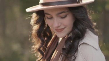 Portrait beautiful girl in nature. Young brunette woman in stylish hat and raincoat is walking along the road. Smiling and happy lady is carefree walking along forest road in spring, autumn or summer
