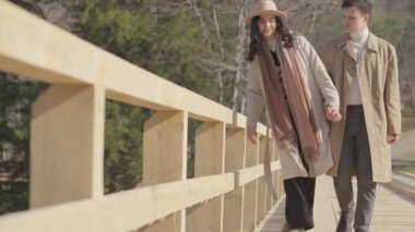 Boy and girl running on wooden bridge over river in mountains. Happy carefree young couple man and woman having fun, romantically and actively resting, joyfully spending time together in relationship.