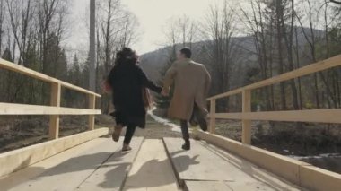 Boy and girl running on wooden bridge over river in mountains. Happy carefree young couple man and woman having fun, romantically and actively resting, joyfully spending time together in relationship.