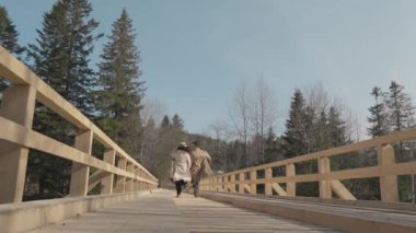 Boy and girl running on wooden bridge over river in mountains. Happy carefree young couple man and woman having fun, romantically and actively resting, joyfully spending time together in relationship.