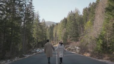 Romantic couple walking along highway in mountains forest. Man takes woman hand and romantic date outdoors. Joyful people boy and girl sit, walk on road. Love couple having fun while walking in nature