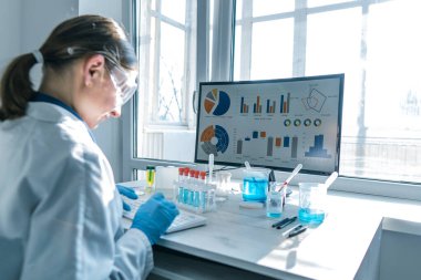 Bir kadın araştırmacı, bir laboratuarda, kişisel bilgisayar monitörünün arkasında oturuyor. Arkaplan araştırma istatistiklerine karşı. Korumada çalışan ilaç uzmanı klavyede çalışıyor..