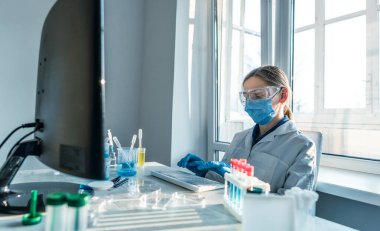Bir kadın araştırmacı, bir laboratuarda, kişisel bilgisayar monitörünün arkasında oturuyor. Arkaplan araştırma istatistiklerine karşı. Korumada çalışan ilaç uzmanı klavyede çalışıyor..