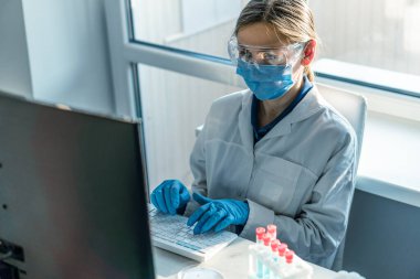 Bir kadın araştırmacı, bir laboratuarda, kişisel bilgisayar monitörünün arkasında oturuyor. Arkaplan araştırma istatistiklerine karşı. Korumada çalışan ilaç uzmanı klavyede çalışıyor..