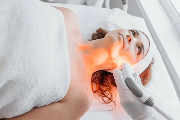 The photo shows the process of light therapy for facial skin rejuvenation in a beauty salon. LED therapy reduces redness and increases hydration of the skin, improving its texture and tone. High