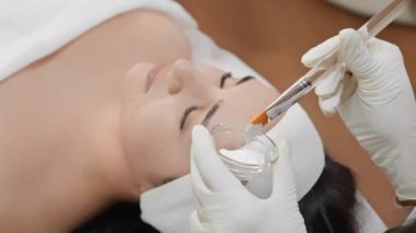 Güzellik uzmanı bir güzellik salonuna kozmetik maske takarak bir kadının yüzünde sihir yaratıyor. Güzellik ve özenin vücut bulmuş hali videosu. Genç bir kadın spa tedavisinden hoşlanıyor. Yenileme ve