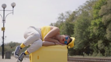 Spor ayakkabılı ve şapkalı bir kız bavulunda yan yatar. Bir kadın yaz mevsiminde tren istasyonunda bir tren beklerken valizinin üzerinde dinleniyor. Tatil ve denize yapılacak bir geziyi bekliyorum. Yüksek