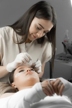 A woman is relaxed at the procedure at a beautician in a beauty salon, and the lashmaker carefully extends her eyelashes, creating a wonderful decoration effect for her face and emphasizing her beauty clipart