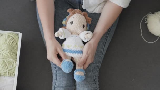 女性の手は自分の足にニット人形を保持します 糸で作られた柔らかいおもちゃを熱心に編み上げ 糸をほどき 赤ちゃんの心を暖めるユニークなディテールを作り出します 高品質4K — ストック動画