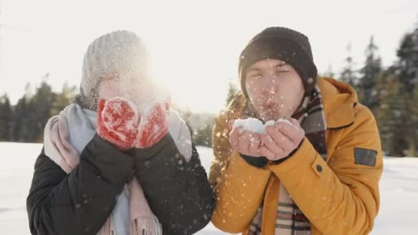 恋人の若い幸せなカップルは冬に彼らの手のひらからカメラに雪を吹く 男と女は冬の気分とクリスマスと新しいの休日の魔法のおとぎ話の期待を与える — ストック動画