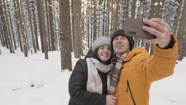 冬の森の中で冬の散歩を楽しんでいる幸せなカップル 若いカップルは美しい自然をキャプチャするためにスマートフォンで自撮りを取ります 男と女が一緒に撮影される — ストック動画