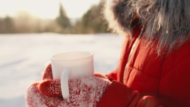 在大自然的冬天喝一杯热饮 一个穿红衣服的女人拿着她戴着手套的手 拿着一杯热饮 然后她就把它蒸了出来 圣诞节和新年快到了 假期和放松 — 图库视频影像