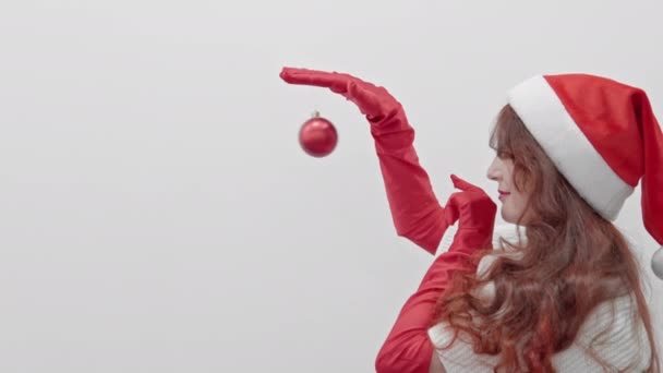 一个在白色背景上戴着圣诞帽的女人在玩圣诞舞会 一个戴着红色手套的女孩拿着一个红色圣诞树玩具 除夕之夜红头发精灵 圣诞节的冬天气氛 — 图库视频影像