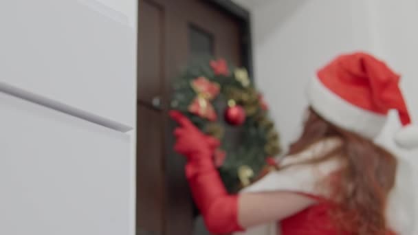 一个穿着红色衣服 头戴圣诞帽的迷人女人手里拿着一个新年花环 紧贴着她家的前门 圣诞节气氛很好 假期快到了 家庭装修 — 图库视频影像